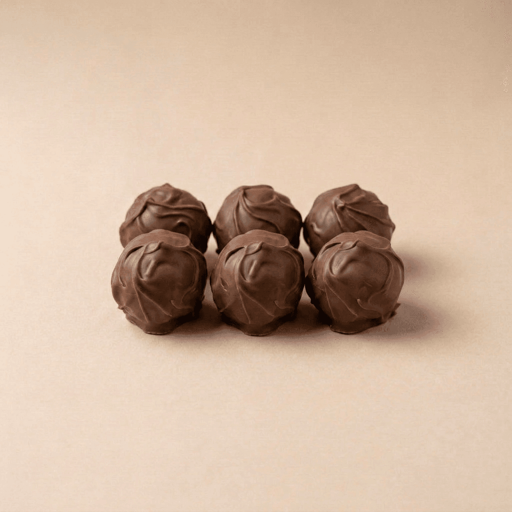 Zes ronde ChocoDough-balletjes met een omhulsel van romige melkchocolade, stijlvol gerangschikt op een zachte beige achtergrond. De chocolade heeft een ambachtelijke, golvende afwerking die de smeuïge en luxe uitstraling benadrukt.