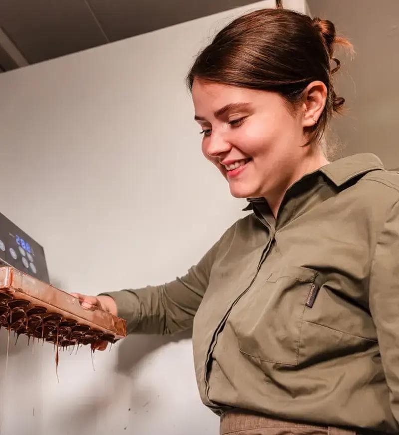 Een glimlachende persoon houdt een chocoladevorm of bakplaat vast terwijl gesmolten chocolade langs de onderkant druipt. De persoon draagt een olijfgroen overhemd en staat binnen tegen een lichte muur.