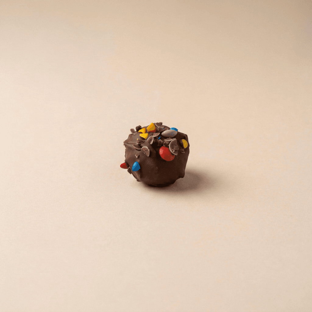 Eén Rainbow Choco Dough-bal met een romige melkchocoladecoating, afgewerkt met kleurrijke smarties en chocoladeschilfers, gepresenteerd op een zachte beige achtergrond. De topping zorgt voor een speelse en knapperige uitstraling.