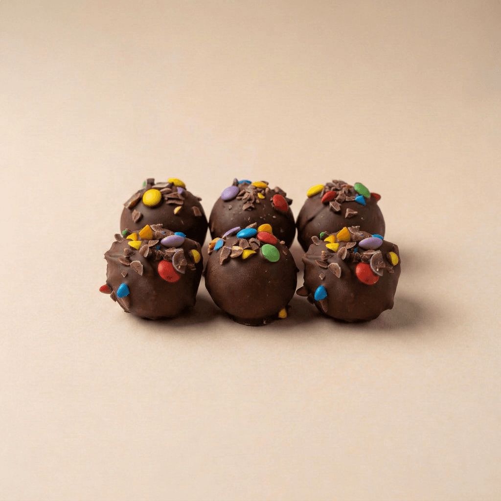 Zes Rainbow Choco Dough-ballen met een romige melkchocoladecoating, afgewerkt met kleurrijke smarties en chocoladeschilfers, stijlvol gerangschikt op een neutrale beige achtergrond. De combinatie van chocolade en kleurrijke toppings geeft een feestelijke en luxe uitstraling.